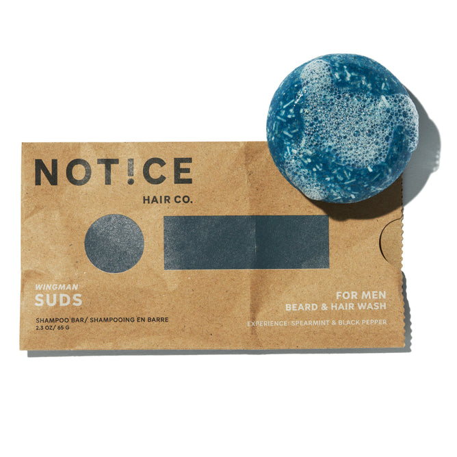 The Suds Shampoo Bar by NOT!CE Hair Co is a blue shampoo bar with white suds on a brown package. Designed for men, it washes beard and hair with spearmint and black pepper fragrance, promoting sustainable, plastic-free self-care.