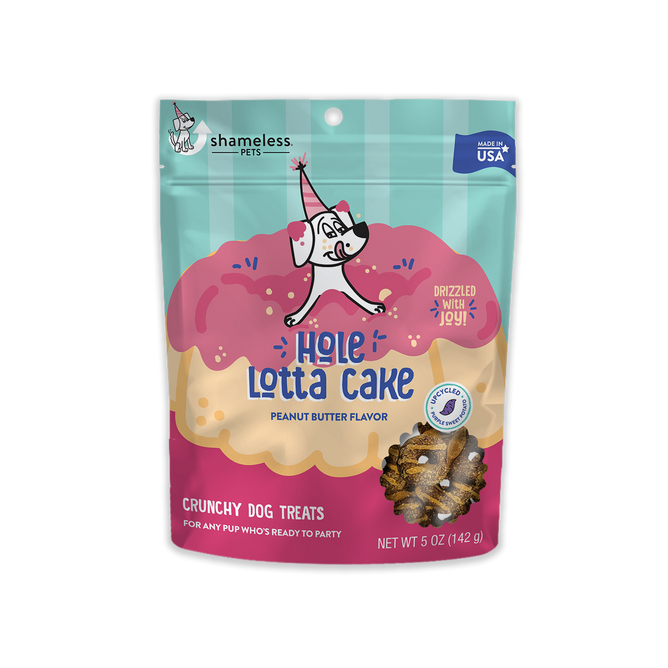 A colorful package of Shameless Pets Hole Lotta Cake - Crunchy Dog Treat in peanut butter flavor features upcycled sweet potatoes. The cartoon dog with a party hat sits on a pink frosted cake, and the bags weight is 5 oz (142 g).