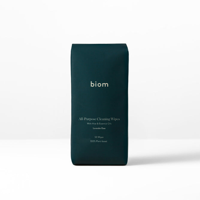 The upright dark green package on the white surface, labeled Biom, contains All-Purpose Wipe Refills. These wipes are infused with aloe, essential oils, and soothing lavender, made from naturally derived ingredients. The convenient pack of 50 wipes is 100% plant-based.