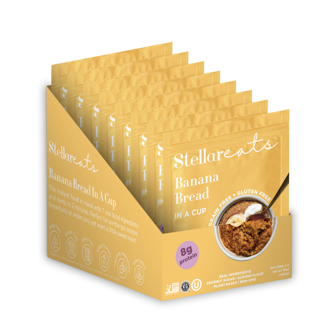 A display box holds several yellow packets of Stellar Eats Banana Bread In A Cup, highlighting features such as grain-free, gluten-free, and 8g protein. Ideal for paleo-friendly diets, each packet displays an image of instant mug cakes.