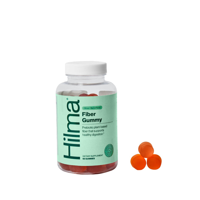 A green-labeled bottle of Hilmas Fiber Gummy displays Prebiotic plant-based fiber for gut health. Nearby are three orange gummies.