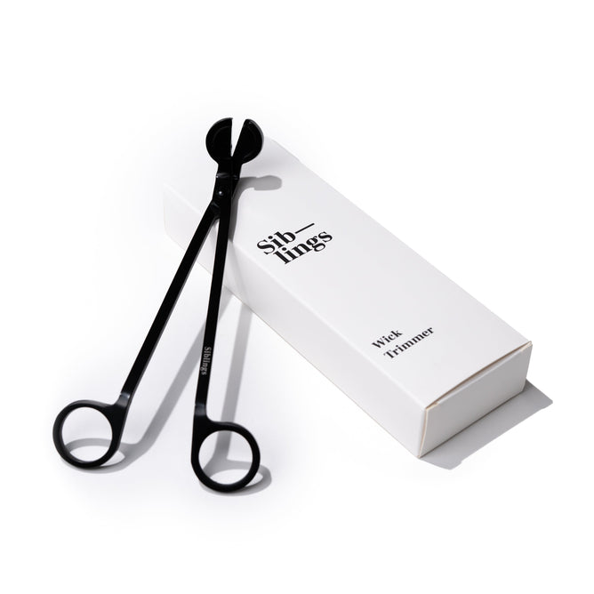 The Wick Trimmer by Siblings boasts a sleek matte black finish, elegantly resting on a white box labeled Siblings Wick Trimmer with refined Siblings engraving against a pristine white background.