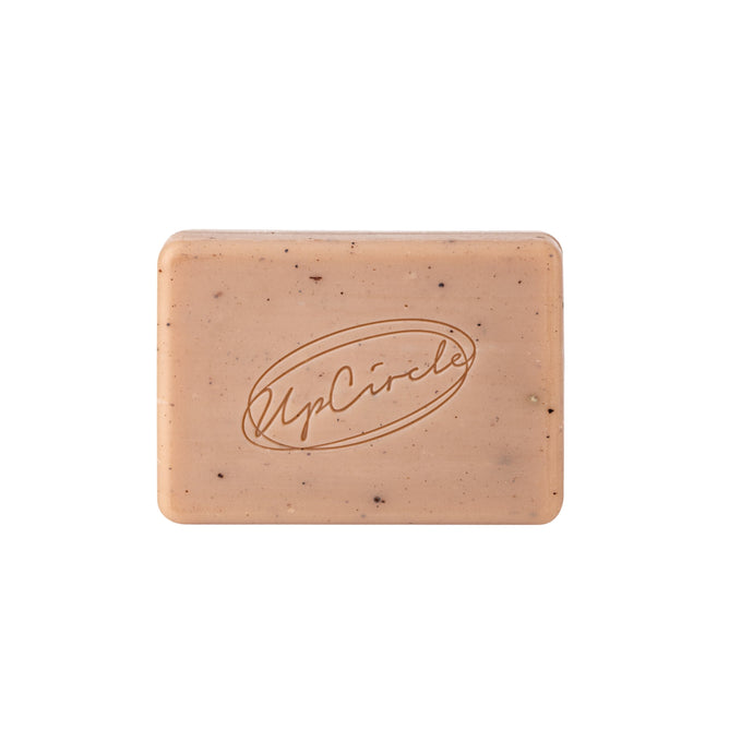 The UpCircle Cleansing Soap Bar with Cinnamon + Ginger is a rectangular, light brown soap engraved with the UpCircle logo, featuring speckles and rounded edges, infused with pink clay for gentle exfoliation and a radiant complexion.