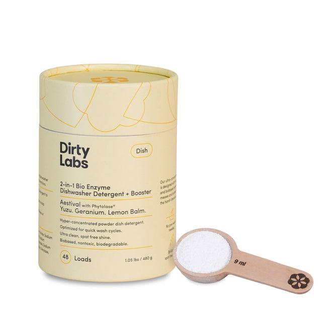 A yellow-labeled cylindrical container of Dirty Labs 2-in-1 Bio Enzyme Dishwasher Detergent + Booster stands beside a wooden measuring spoon with white powder, promising biobased, bioenzyme-powered cleaning for 48 loads and sparkling results.