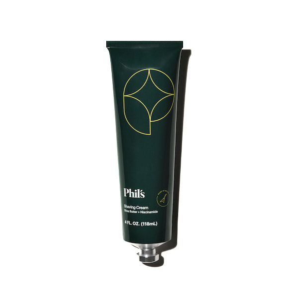 A dark green tube features a minimalist yellow geometric logo with Phils Shaving Cream in white. Enhanced with shea butter, it holds 4 fl oz (118 mL). A silver cap complements the design, and the tube casts a shadow on the right.