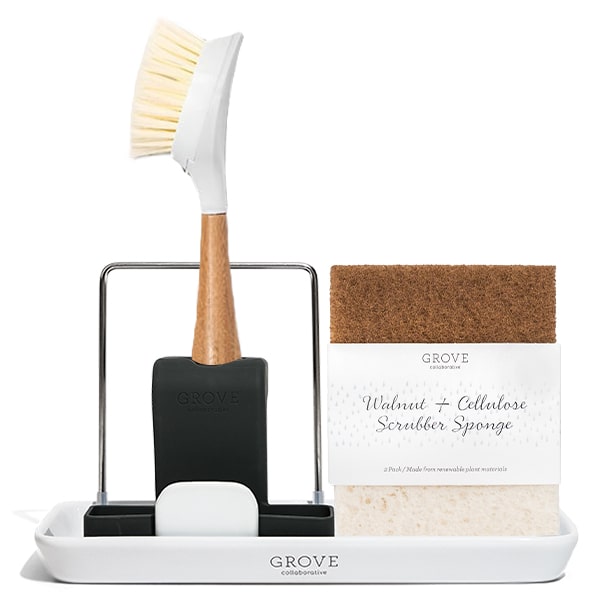 A wooden-handled dish brush is in a black holder on a white Grove Collaborative tray. Next to it, theres a pack of Grove Collaborative Walnut + Cellulose Scrubber Sponge, featuring a brown scrub side and white sponge side. This setup is part of the sinkside-tool-set by Grove Collaborative.