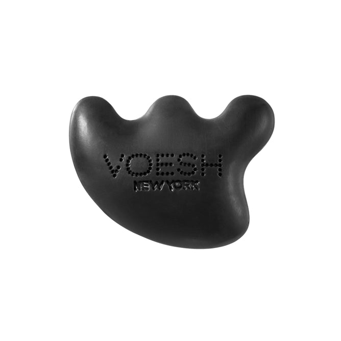 The Gua Sha Cleansing Bar by VOESH New York is a black, pebble-shaped massage tool with VOESH NEW YORK formed by small holes. It features three rounded bumps and is infused with detoxifying charcoal powder for an ergonomic, firm grip suited for effective massage therapy.