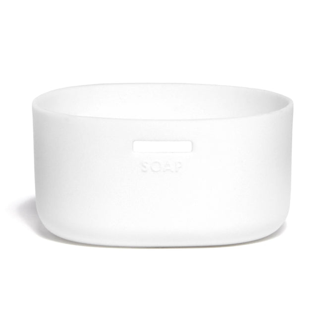 A white, oval soap dish embossed with SOAP stands out with its minimalistic design and matte finish. Nearby, a Grove Co. Silicone Sleeve Glass Foaming Hand Soap Dispenser adds elegance, complementing the simplicity against the pristine background.