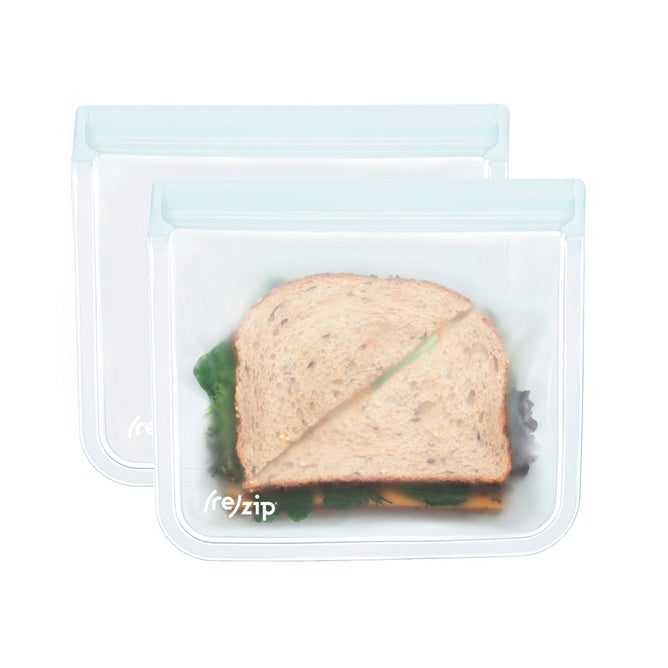 Two Flat Lunch Bags by (re)zip, crafted from transparent silicone, display sandwiches with whole grain bread, visible lettuce, and cheese. The airtight, leakproof bags are sealed at the top and set against a plain white background.