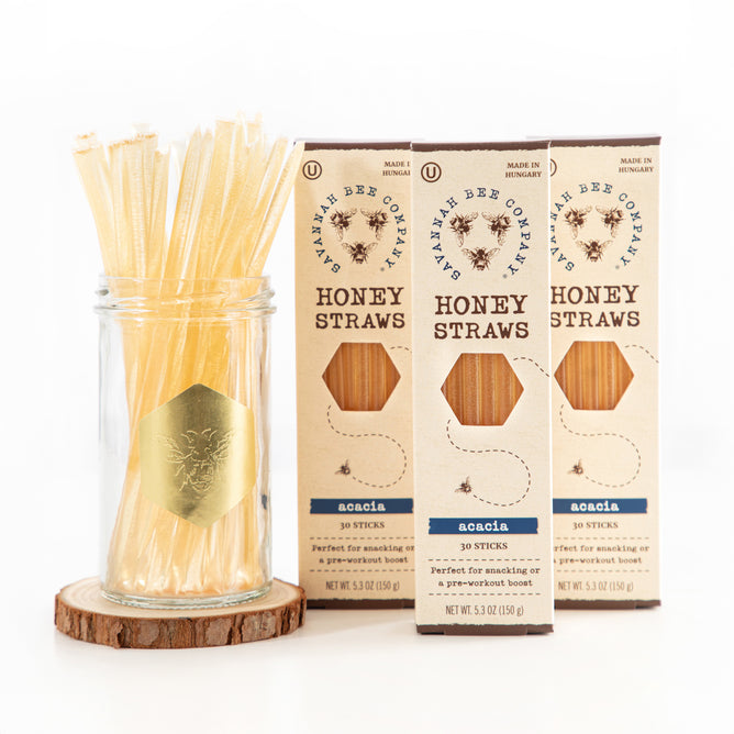 A glass jar filled with Acacia honey straws, wrapped in BPA-free plastic and featuring a bee logo, sits next to three elegant boxes of Savannah Bee Companys Honey Straws. These boxes emphasize the natural charm and sophisticated presentation of their Acacia flavor.