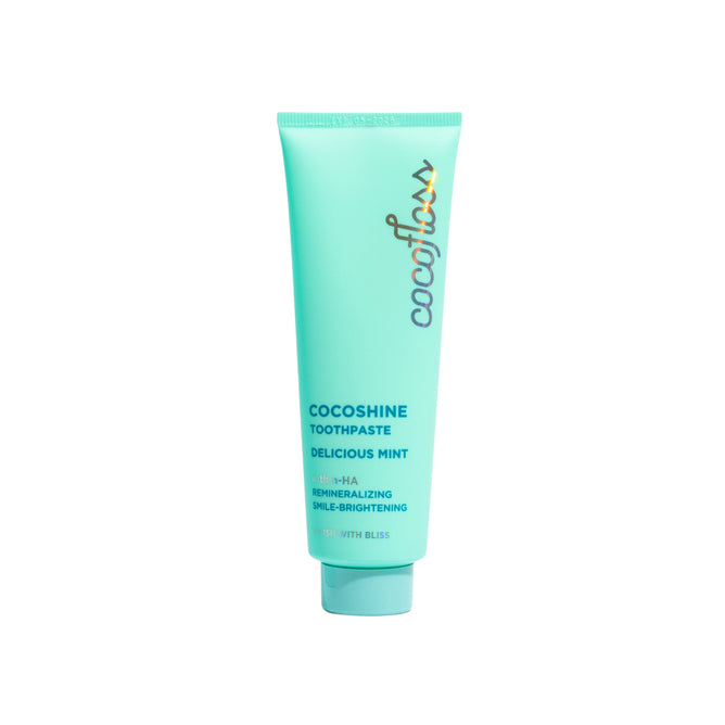 The turquoise tube of Cocofloss Cocoshine Whitening Toothpaste in Delicious Mint flavor enhances enamel remineralization and smile brightness with nano-hydroxyapatite. The packaging features a colorful brand logo and white lettering.