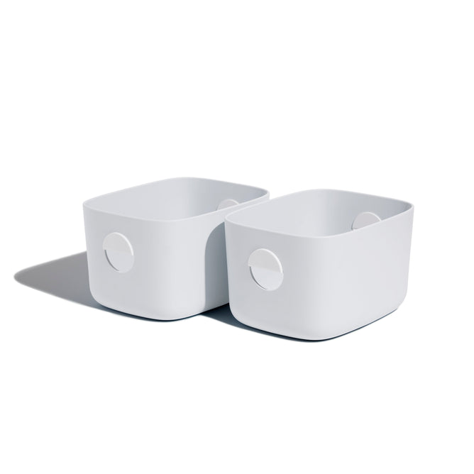 Two Open Spaces Medium Storage Bins, featuring rounded corners and circular handles, are neatly placed side by side on a shelf against a white background. They cast soft shadows to the left, adding an elegant touch of multi-purpose storage.