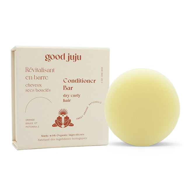 A round, beige conditioner bar sits beside its box labeled in French and English as the Dry/Curly Conditioner Bar. Infused with sweet orange and patchouli, it tames frizz using organic ingredients. The brand name Good Juju Body & Home adorns the box.