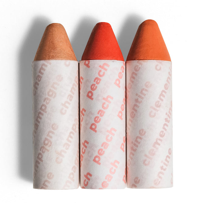 The Axiology Balmie Trio features three crayon-like, vegan, multi-use lip-to-lid balms standing vertically: a beige one labeled Champagne, a red one labeled Peach, and an orange one labeled Clementine. Each has a white wrapper with the color name in matching text.