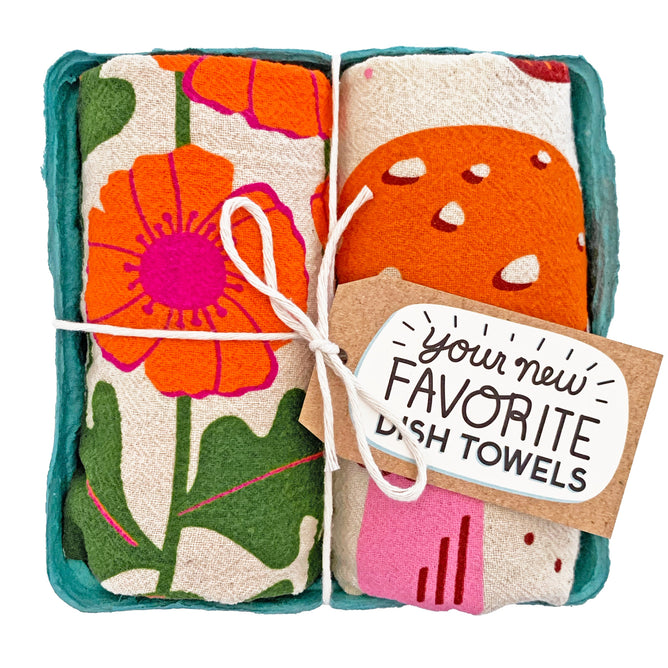 A set of two super absorbent dish towels from The Neighborgoods features colorful floral and mushroom designs. Made of 100% unbleached cotton, they come tied with twine and a tag saying Your New Favorite Dish Towels, elegantly displayed in a teal, rectangular holder.
