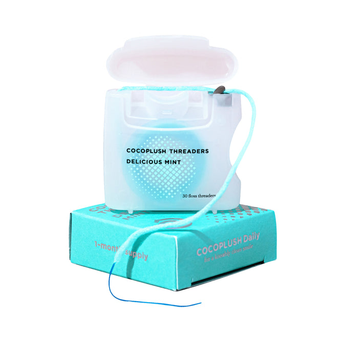 An open plastic container of mint-flavored floss-and-threader combo labeled Cocofloss Threaders Delicious Mint, with a strand extending out, sits atop a teal box marked 1-month supply Cocofloss Daily.