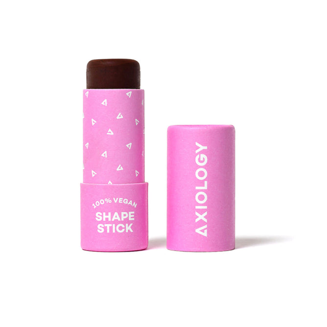 An open pink Axiology Shape Stick reveals a brown contour inside. The packaging features white text and triangle patterns, declaring 100% Vegan.
