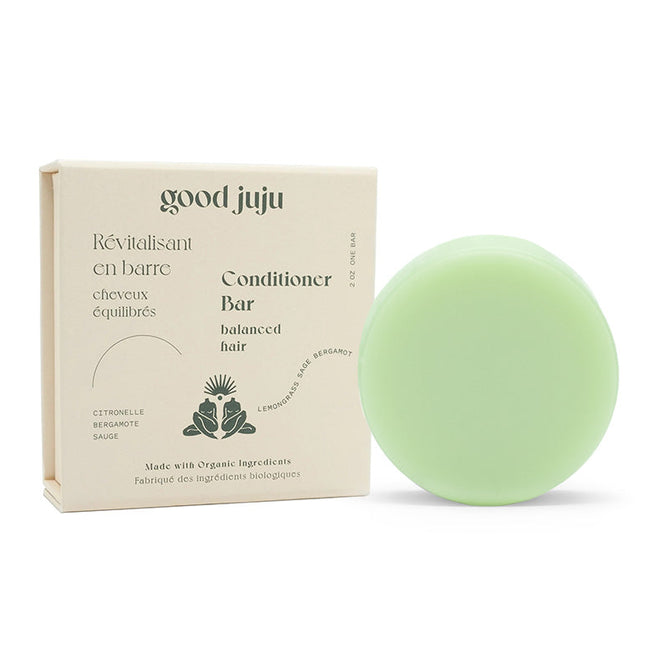 A green, round conditioner bar and a beige box marked Good Juju Body & Home contain the Normal/Balanced Conditioner Bar, made with organic ingredients such as lemongrass, bergamot, sage, and jojoba oil. The packaging includes French and English text for balanced hair care.