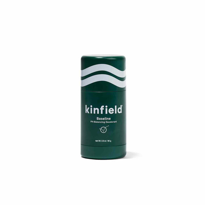 A Kinfield Baseline Deodorant stick, featuring a green hue with wave designs and text, stands upright against a white backdrop. This eco-friendly product is infused with pH-balancing fruit enzymes and offers a recyclable refill option.