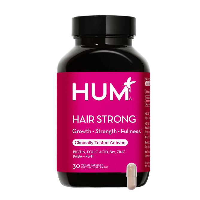 A black bottle with a bright pink label shows the brand HUM Nutrition and Hair Strong - Capsules for Growth with Biotin, Fo-Ti, & Paba. Its a dietary supplement aimed at boosting hair growth with benefits like strength and fullness emphasized. A single beige capsule is placed in front.