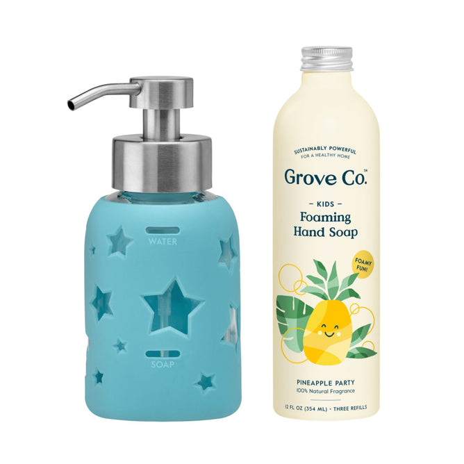 A blue foaming soap dispenser with star and moon cutouts is paired with a bottle from the Grove Co. Kids Foaming Hand Soap Starter Set, decorated with a smiling pineapple and labeled Pineapple Party, emphasizing its natural scent and plastic-free packaging.