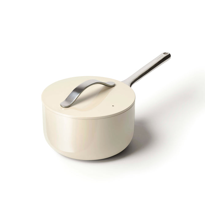 The Caraway Ceramic Sauce Pan (3 qt) is beige with a ceramic coating, non-toxic design, and a long stainless steel handle. It has a matching lid featuring a stylish, curved handle for easy lifting, complementing the elegant design on a white background.