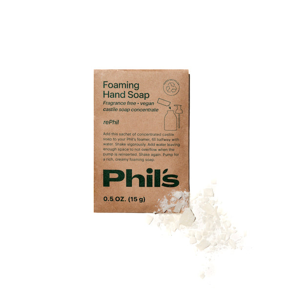 A small brown cardboard sachet, labeled Hand Soap RePhils by Phils, sits beside scattered soap flakes. This fragrance-free, vegan formula is crafted with organic castile soap concentrate and packaged in a recyclable paper envelope.