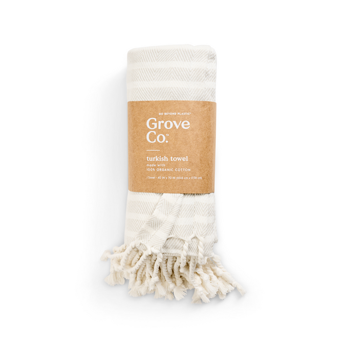 A folded ivory Turkish towel with beige stripes, labeled Grove Co. from the Spring Collection, features an organic cotton tag. It has tasseled edges and is wrapped in a brown eco-friendly paper band emphasizing its sustainable commitment.