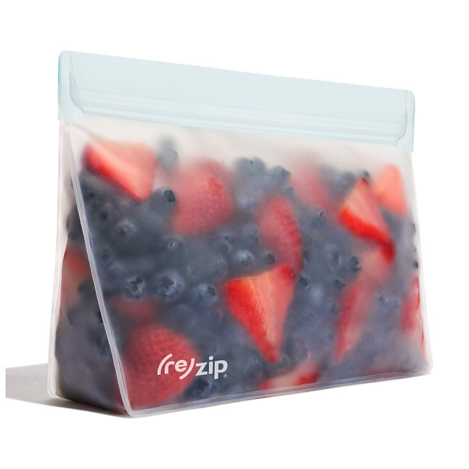 A Stand-Up Quart - 4 Cup Bag by (re)zip, made of food-safe PEVA, showcases blueberries and sliced strawberries. This translucent, reusable bag features a light blue, leakproof airtight closure and stands upright to beautifully display fresh fruit.