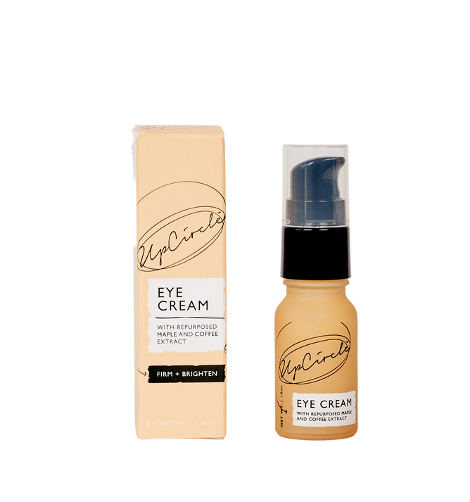 A pump bottle of UpCircles Eye Cream with Cucumber, Hyaluronic Acid, and Coffee sits next to its box. The label highlights Repurposed maple and coffee extract. Firm + Brighten. Designed for dark circles, the light brown packaging features sleek black text.