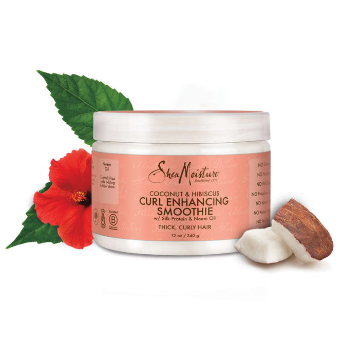 A jar of SheaMoisture Coconut + Hibiscus Curl Enhancing Smoothie rests on a white backdrop, accompanied by a hibiscus flower, neem leaves, and coconut. The anti-frizz formula features nourishing ingredients for thick, curly hair.