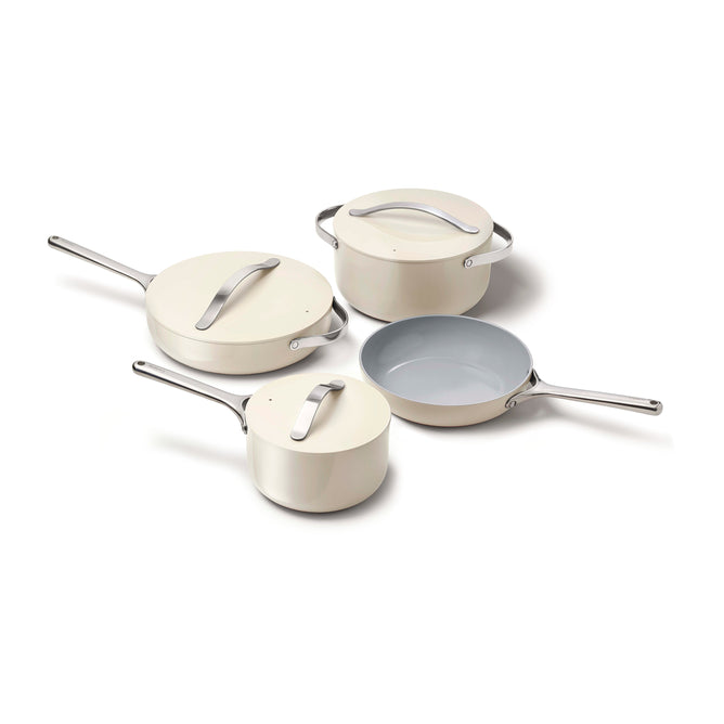 Four beige pieces from the Caraway Nonstick Ceramic Cookware Set lie elegantly on a white surface, featuring a frying pan with a light blue nonstick interior, two pots with lids, and a saucepan with lid—all adorned with silver handles.