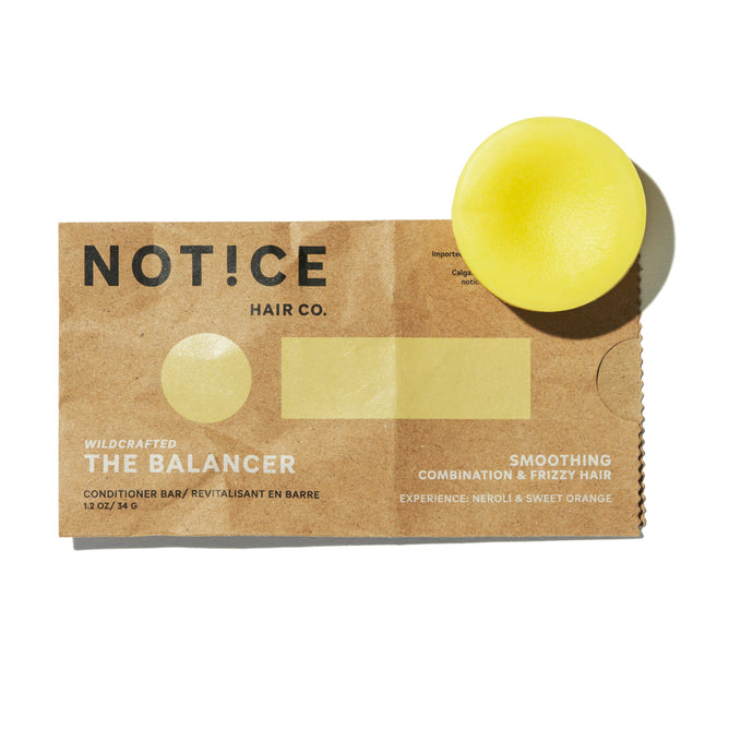 A round, yellow conditioner bar rests on brown paper packaging labeled NOT!CE Hair Co The Balancer. This plastic-free conditioner bar smooths combination and frizzy hair, with a refreshing neroli and sweet orange scent.