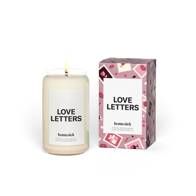 The Homesick Love Letters Candle, a lit white candle in a glass jar, is positioned beside its decorative box adorned with a pink and red stamp pattern. The brand name appears on both the candle and the box.
