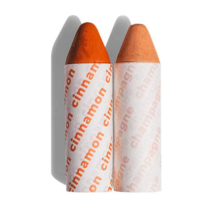 Two Balmie Duo crayons by Axiology stand upright on a white background. The left, cinnamon, has a reddish-brown tip; the right, champagne, features a peach-toned tip. Shadows are cast to the left.
