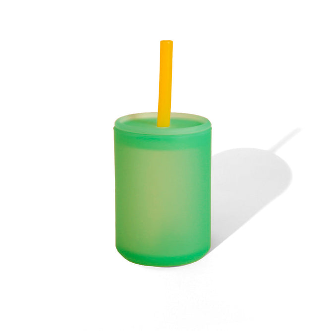 A green Avanchy Training See-Thru Sippy Cup for 6+ months, featuring a lid and yellow straw, stands against a white background, casting a shadow to the right.