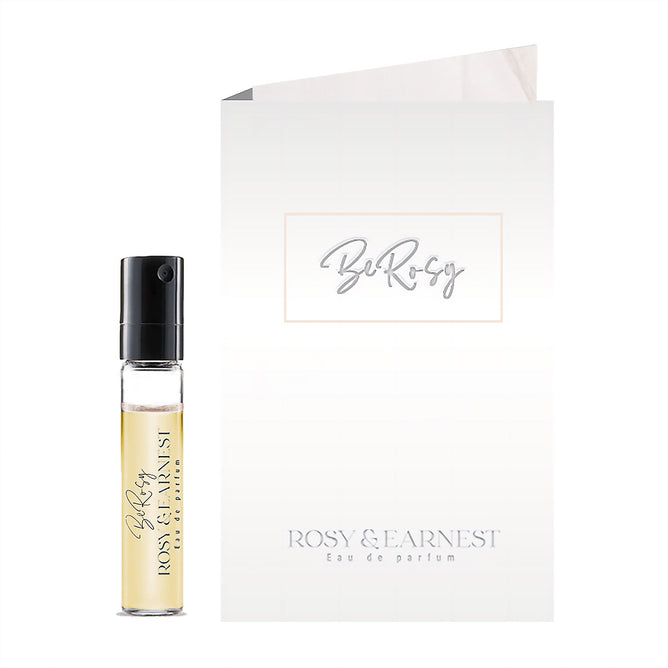 A small sample vial of Be Rosy, eau de parfum by Rosy & Earnest stands before a white card bearing its name, highlighting this vegan fragrance made for mindful beauty lovers.