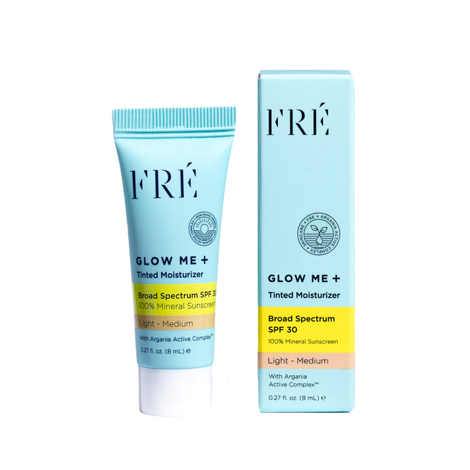 A tube and box of FRÉ Skincare GLOW ME+ Tinted Mineral SPF 30 Deluxe - Sample, light-medium shade, with argania active complex and a non-comedogenic formula, are displayed upright on a white background.