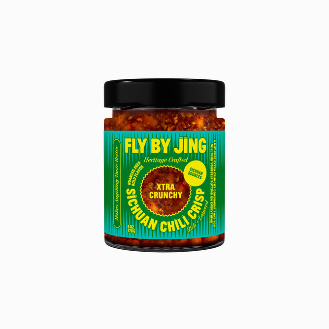 A jar of Fly By Jing Xtra Crunchy Sichuan Chili Crisp, featuring a black lid and vibrant blue-green packaging with yellow and red text. This crunchy condiment is packed with crispy bits for a bold, savory snack.