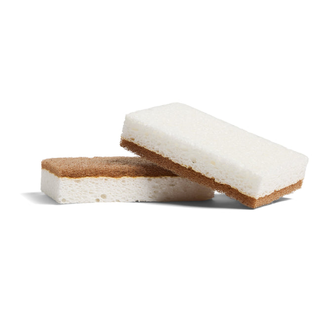 Two Grove Co. Heavy Duty Agave Scrubber Sponges, each with a white soft top and brown abrasive bottom, are shown—one lying flat and the other propped diagonally—against a white background.
