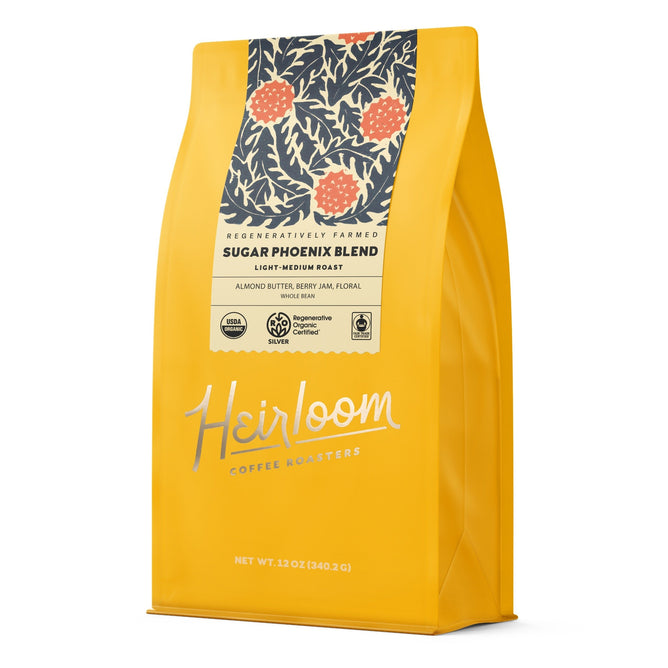 A yellow 12oz bag of Heirloom Coffee Roasters Sugar Phoenix ROC Whole Bean Coffee features a dark label with orange berries and leaves, offering a light-medium roast regenerative organic coffee with almond butter, berry, and floral notes.