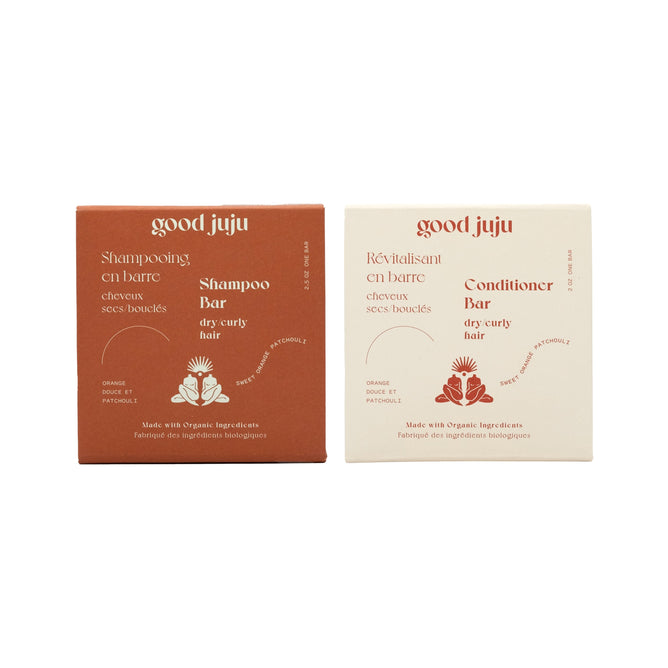 Two square boxes of Good Juju Body & Home’s Dry/Curly Haircare Set are shown on a white background, featuring the rust-colored shampoo bar and cream-colored conditioner bar.