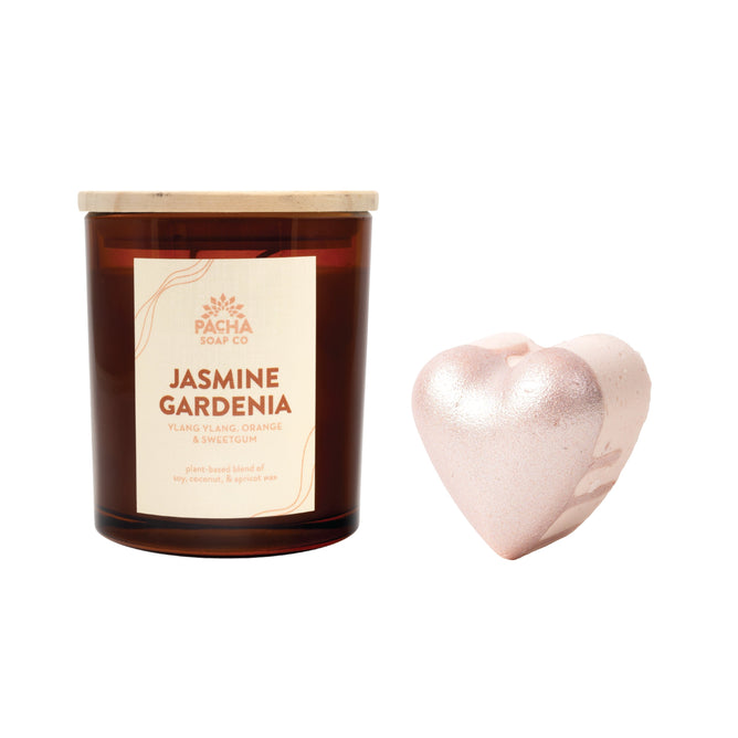 A brown glass Pacha Soap Co. Signature Candle sits beside a shimmery, pink, heart-shaped Jasmine Gardenia Froth Bomb on a white background. Buy a Signature Candle, get the Jasmine Gardenia Froth Bomb free.