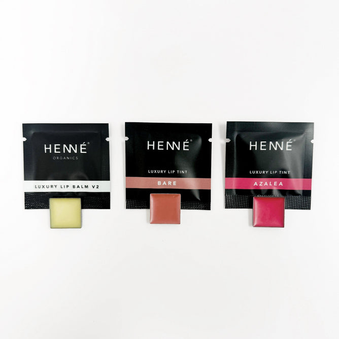 Three Henné Organics Lip Sample Trio products—Luxury Lip Balm V2, Bare Luxury Lip Tint (peachy-pink), and Azalea Luxury Lip Tint (vibrant pink)—are displayed in sleek black packets side by side.