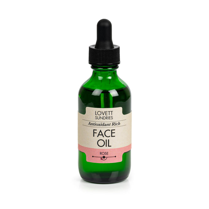 A green glass bottle with a dropper lid labeled Lovett Sundries Face Oil in a rose scent. Enriched with organic oils, it enhances skin elasticity, and the label has a pink bottom section.