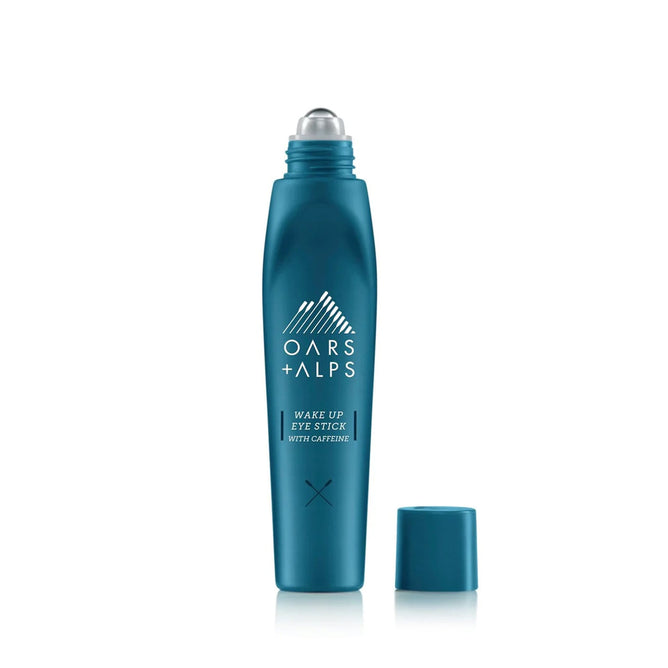 A teal Oars + Alps Wake Up Eye Stick stands upright with its cap beside it, revealing the metal rollerball applicator. This eye serum helps reduce puffiness under the eyes.