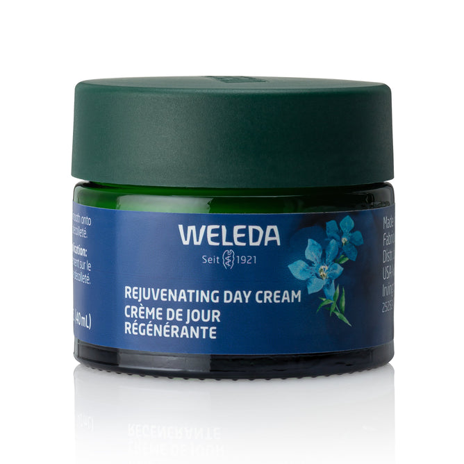 A green jar of Weleda Rejuvenating Day Cream with a dark green lid, featuring blue gentian flower graphics and bilingual English-French text on the label, designed to support mature skin.