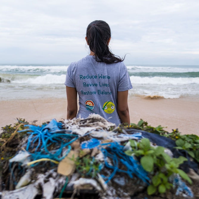 On a sandy beach, a person in a gray shirt with eco messages from Grove Co.s Collect Ocean-Bound Plastic in India stands facing the ocean. Nearby, low-value plastics and debris highlight pollution issues. Waves and cloudy skies emphasize the urgent need for action.