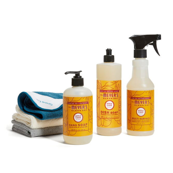The Mrs. Meyers fall-cleaning-set-mmcd, featuring hand soap, dish soap, and multi-surface spray in apple scent, is arranged beside a stack of folded cleaning cloths in white, blue, and gray.