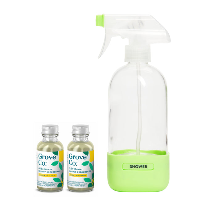 A Grove Collaborative spray bottle labeled SHOWER with a green base stands by two concentrate bottles of the daily shower cleaner, featuring silver caps and green and blue labels.